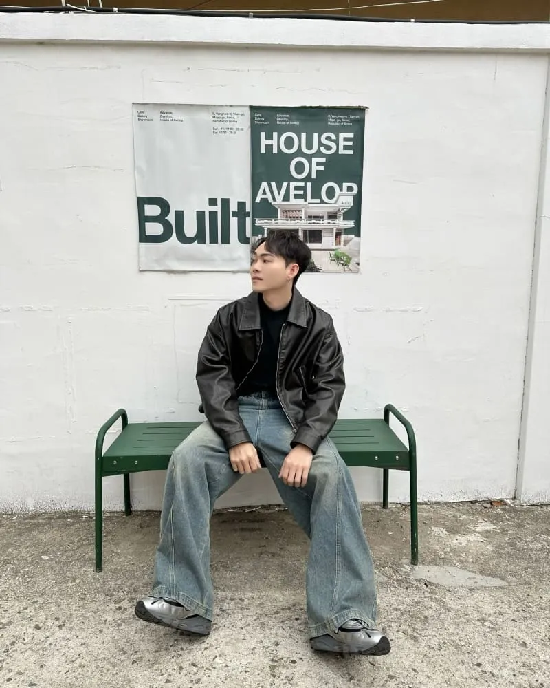 Young man in a black jacket sits on a green bench before a white wall with posters behind him.